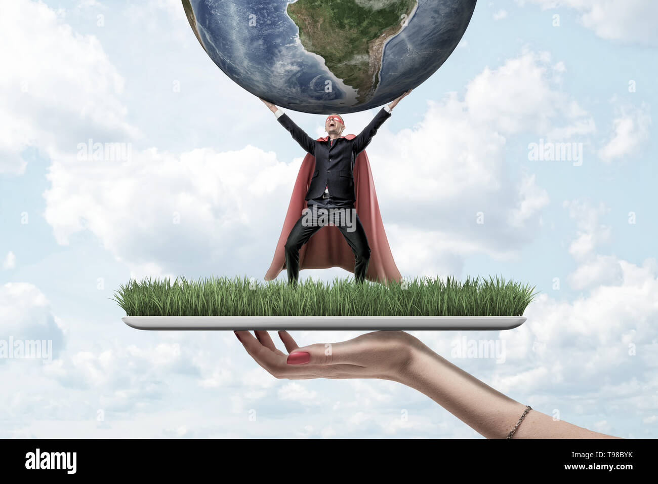 Female hand holding grass board with small businessman wearing red superman mask and cloak holding earth globe up on blue sky background Stock Photo