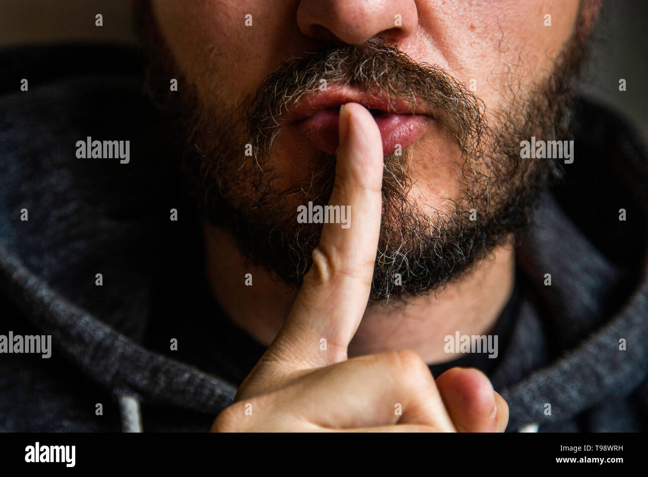 Don't speak, be quiet - close up portrait of bearded man on the door saying shhh. Finger on lips. Head and shoulders, no eyes. Danger. Stock Photo