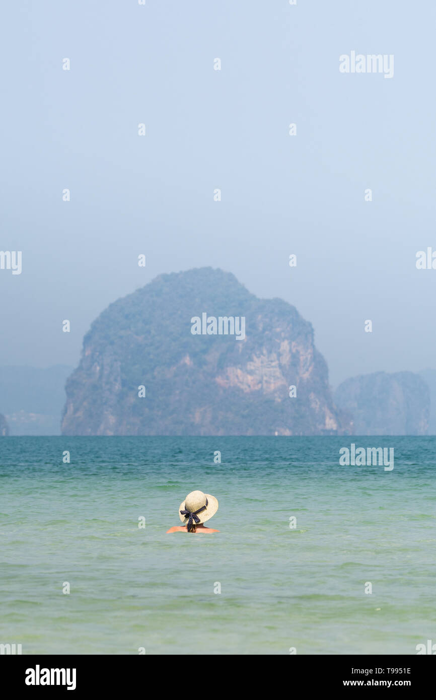 Woman in straw hat swiming in the sea in Krabi Railey beach overlooking the harbour and mountains, Thailand. Vertical orientation Stock Photo