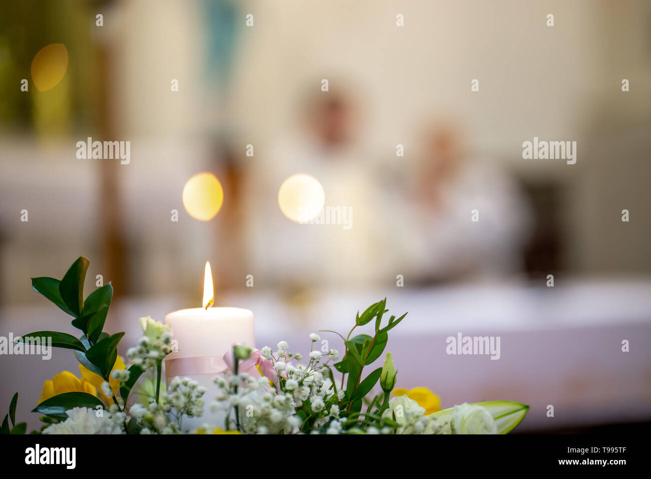 Fragment of burning candle and bouquet of flowers during wedding in church, Latvia. Closeup of wedding flowers bouquet and burning candle on blurred b Stock Photo