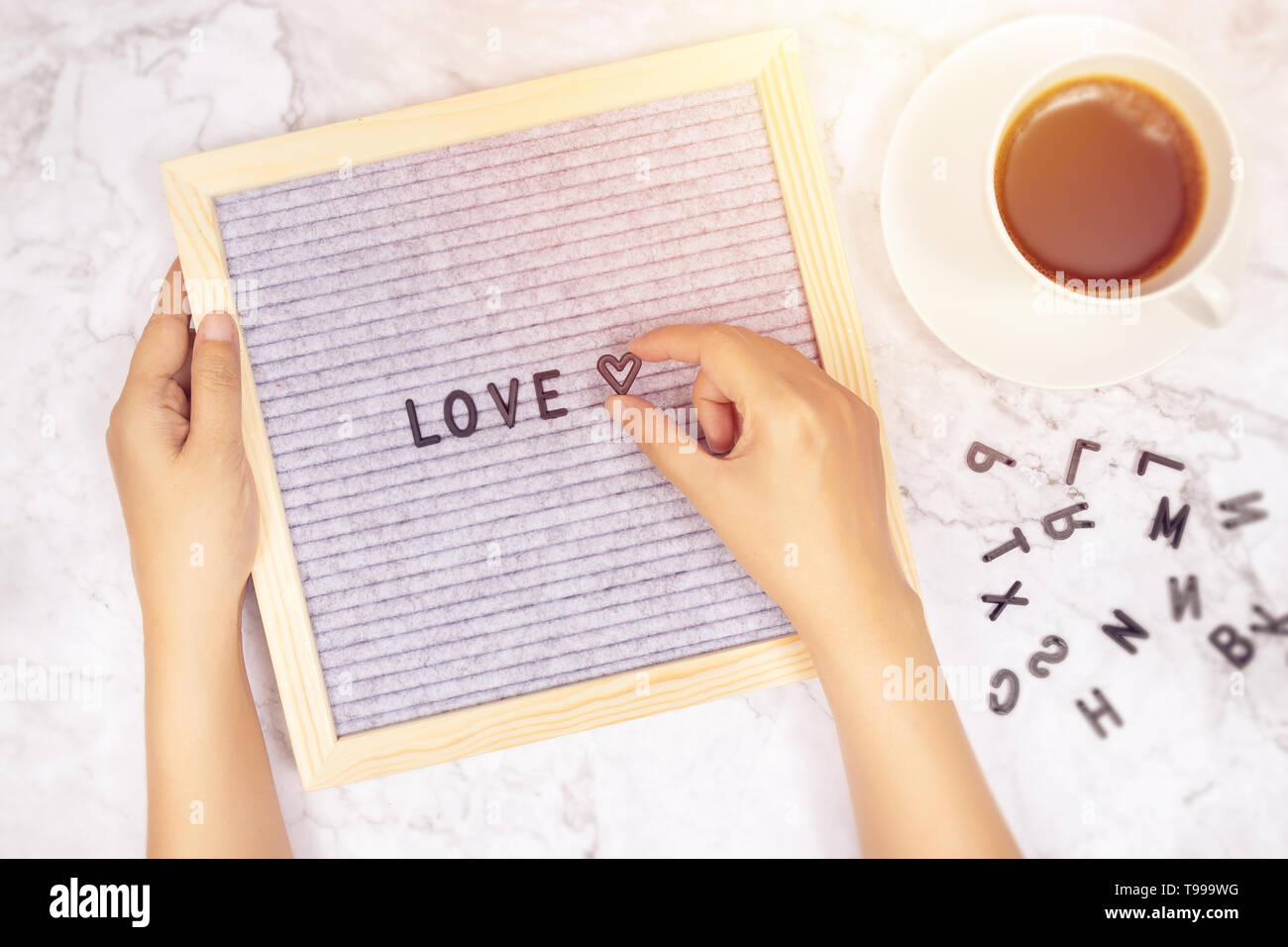 word LOVE on letter board with woman's hand holding heart symbol on white marble desk background with coffee cup Stock Photo