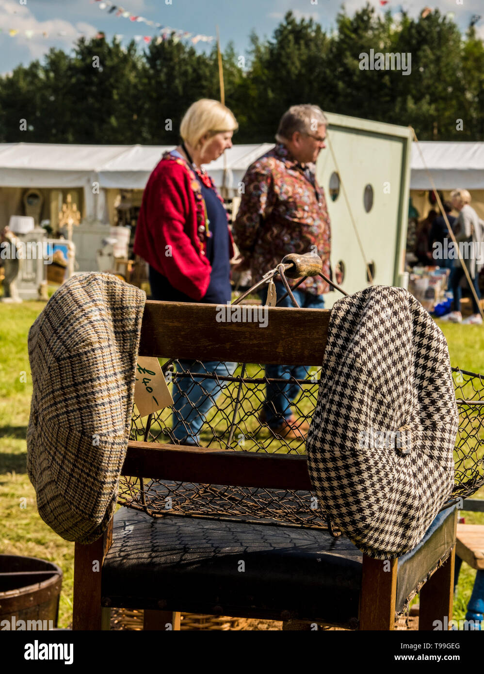 Heterosexual couple walking past antique stall, chair with two flat caps hanging on it in foreground, Ripley, North Yorkshire, England, UK Stock Photo