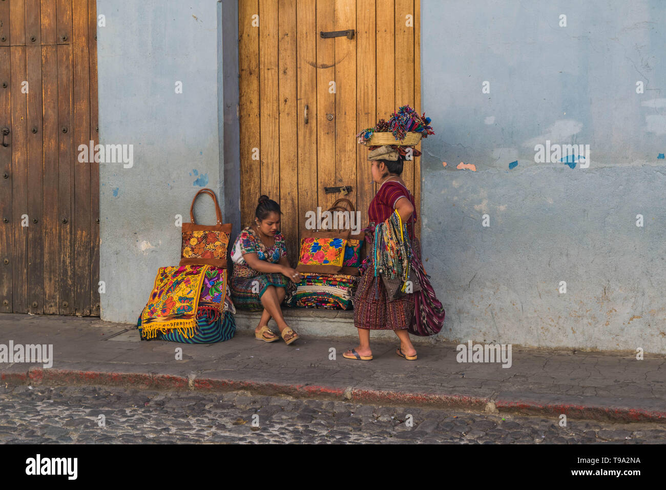 Two women street vendors in traditional clothing, one with a basket on her head, selling Guatemalan goods on a cobblestone street in Antigua Guatemala Stock Photo