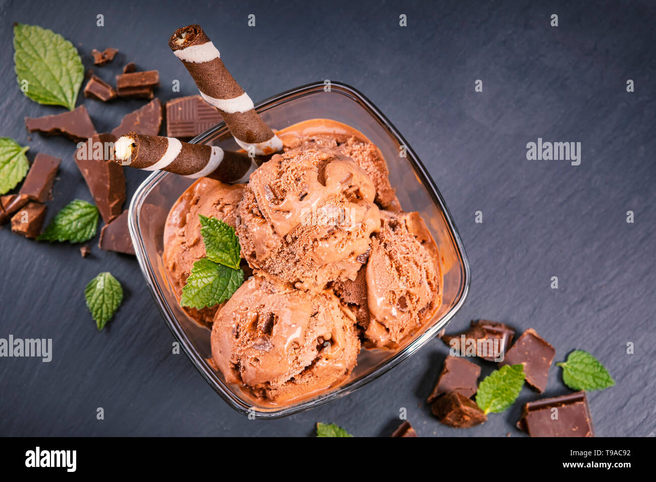 Scoops chocolate ice cream in glass bowl with wafer sticks, cone and chocolate on a black slate board. Focus on Bowl with scoops chocolate ice cream Stock Photo
