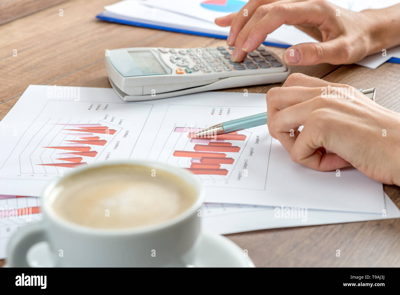 Female accountant working on a presentation or annual report analysing a set of graphs and charts with a manual calculator with a cup of coffee alongs Stock Photo
