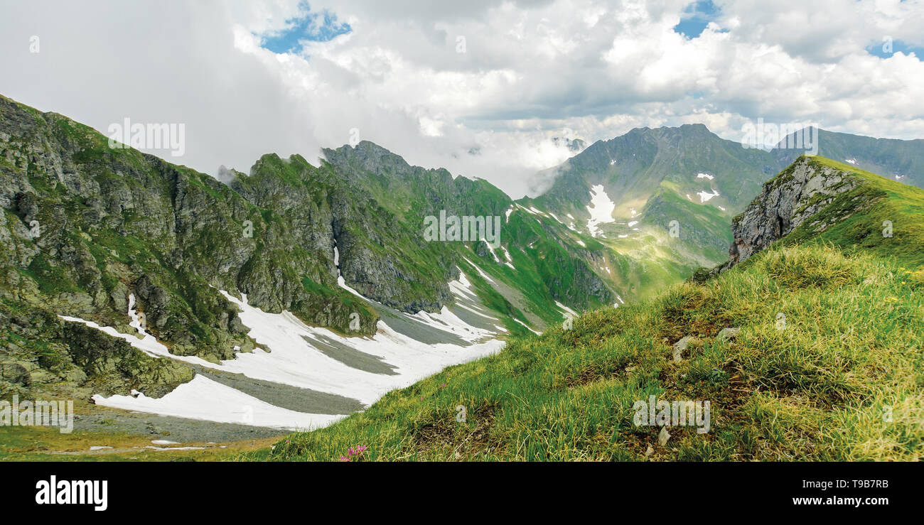 panorama of fagaras mountain ridge in summer. spots of snow on grass of steep slope. rocky tops. cloudy weather. romania landscape Stock Photo