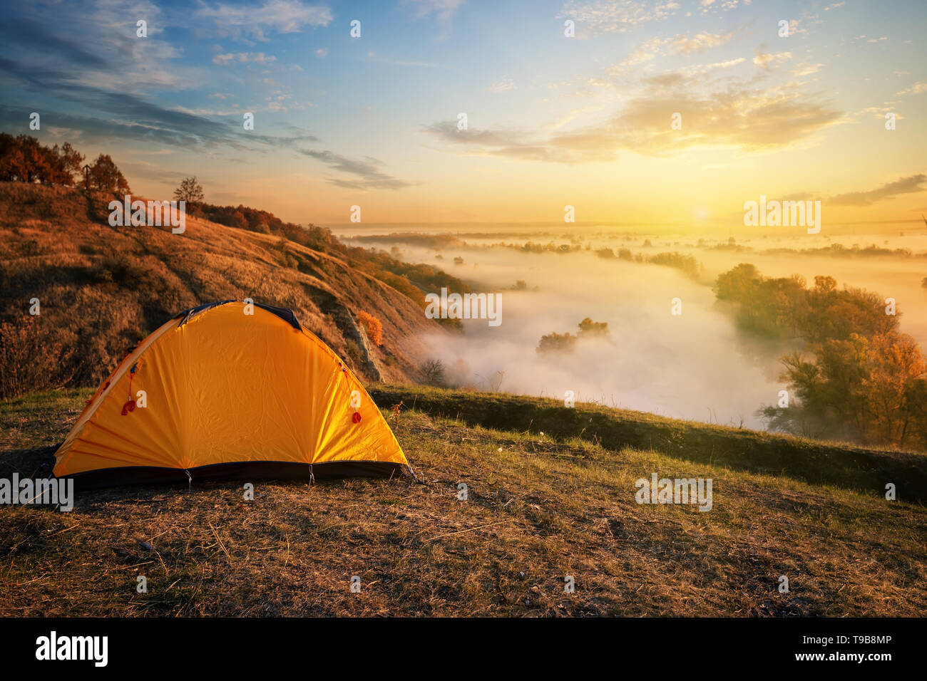 Orange tent in canyon over misty river at sunset Stock Photo