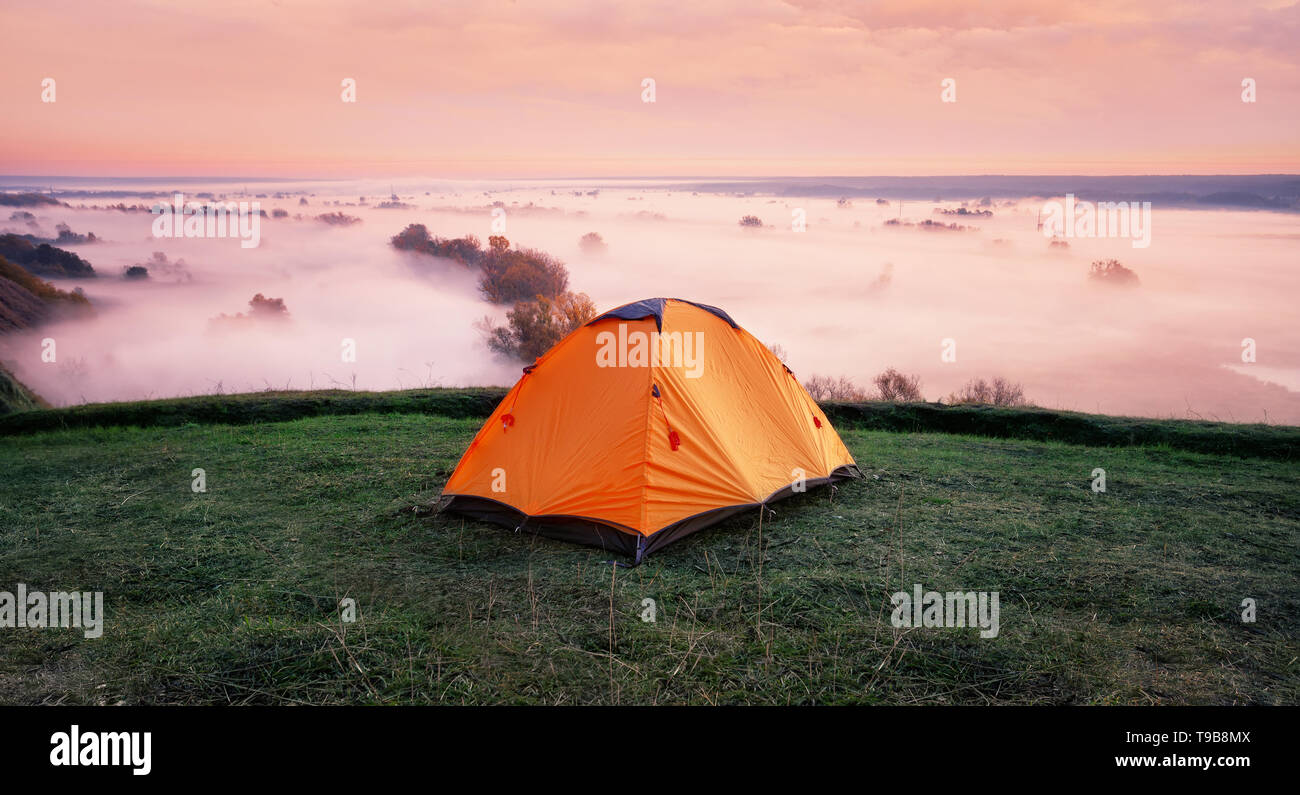 Orange tent on hill above misty river Stock Photo