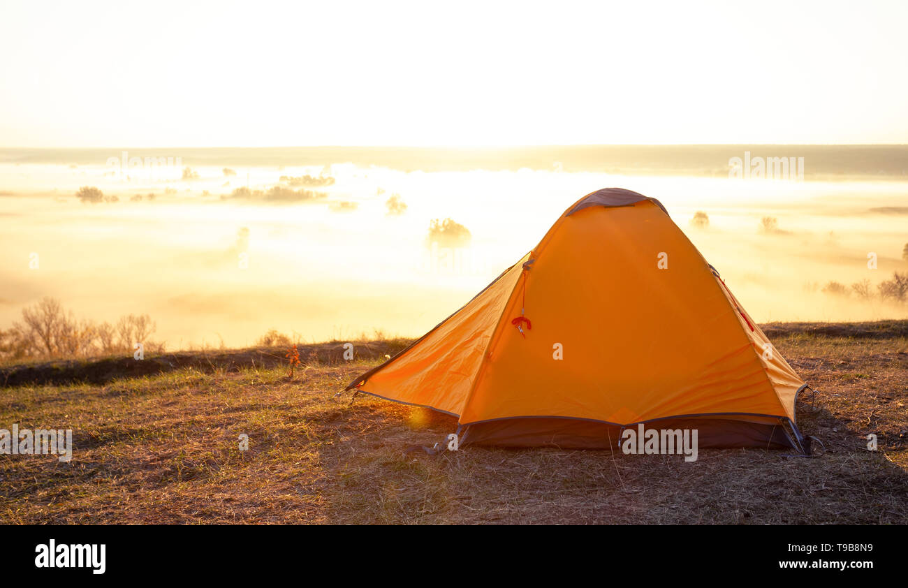 Orange tourist tent on hill above river covered with thick fog Stock Photo
