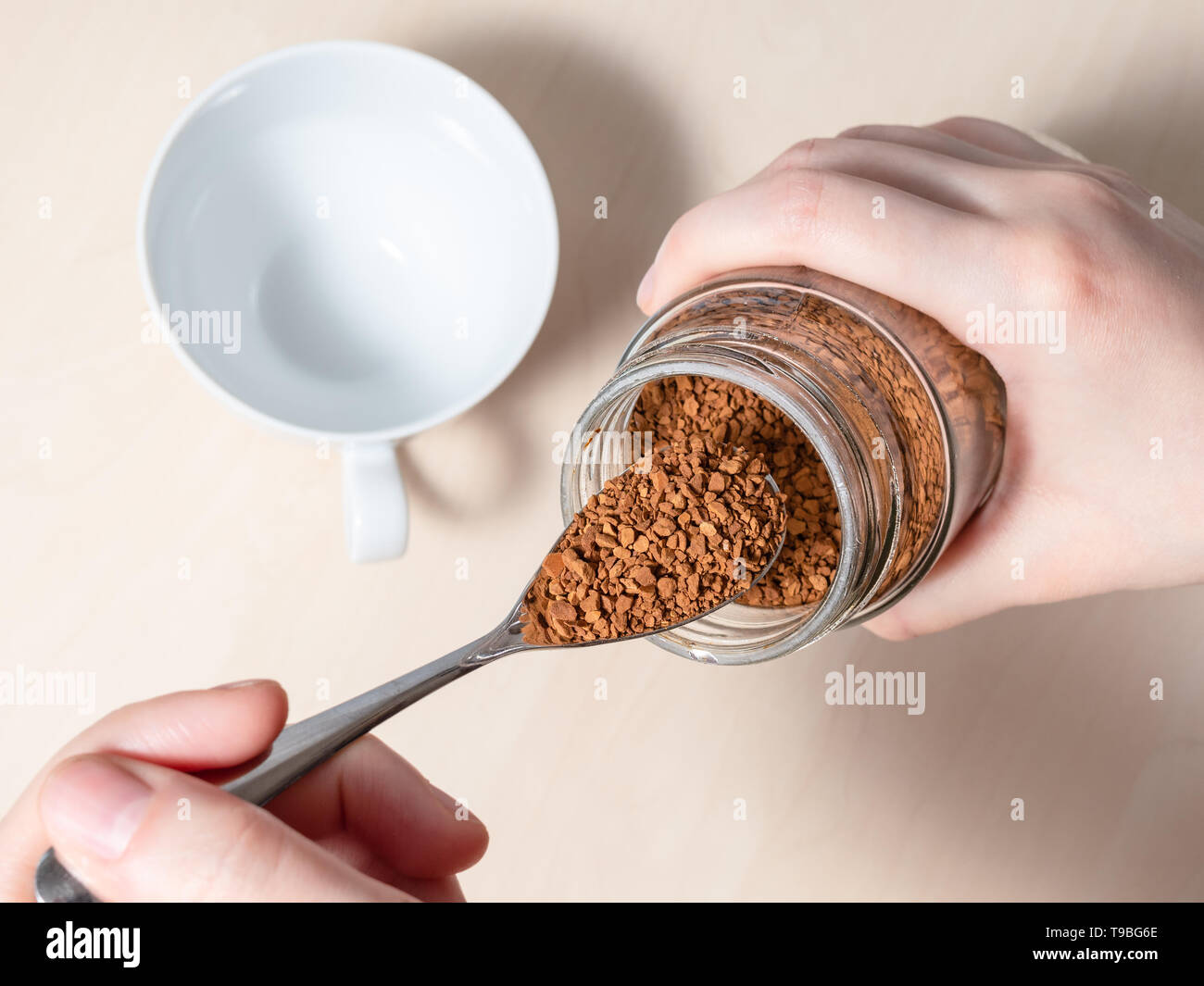 female hand spooning up instant coffee from glass jar over cup on light brown table Stock Photo