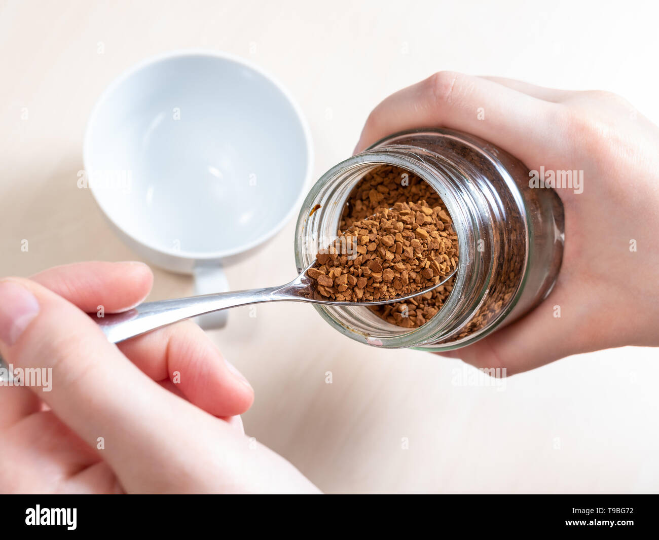 hand spooning up instant coffee from glass jar close up over cup on light brown table Stock Photo