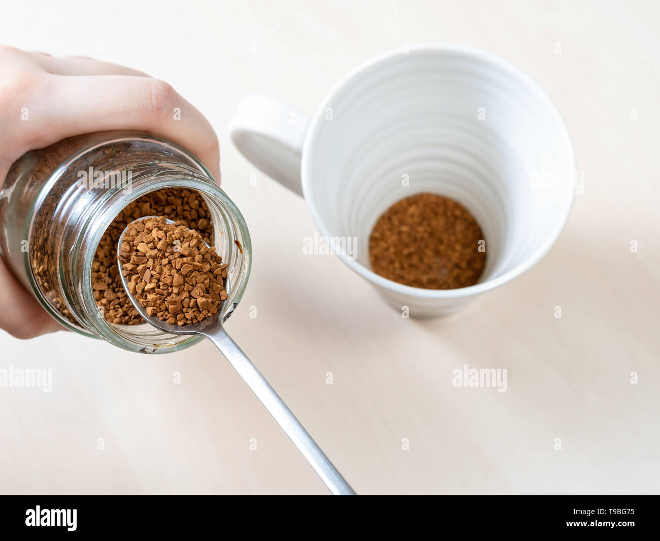 top view spooning up second portion of instant coffee from glass jar over mug on light brown table Stock Photo