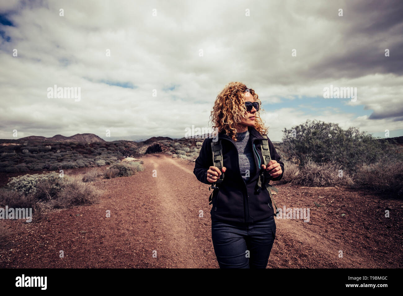 Beautiful trekker caucasian blonde curly young active woman walk in the desert path enjoying the freedom and the active lifestyle traveling with backp Stock Photo