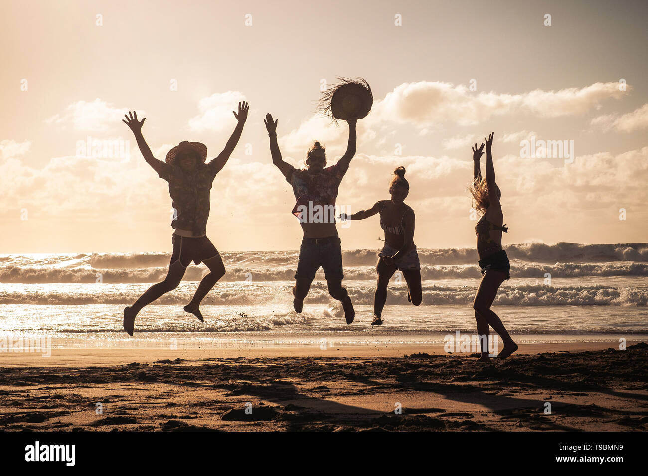 Sunset at the beach with happy group of young people jumping having fun - friends on summer holiday vacation enjoying together in friendship - sandy l Stock Photo