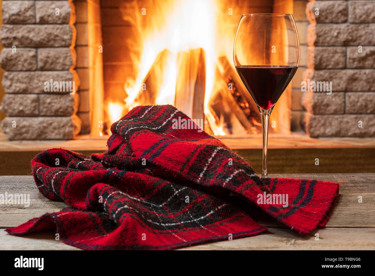 Glass of red wine and warm scarf against cozy fireplace background, in country house, horizontal, hygge , home sweet home. Stock Photo