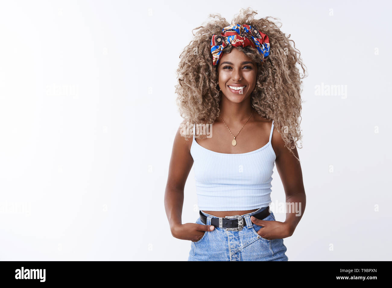 Attractive confident stylish african-american creative woman blond curly hair, white top smiling lively hold hands pockets self-assured determined Stock Photo