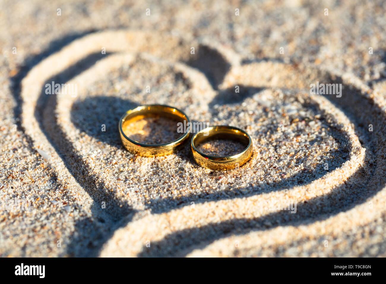 An Overhead View Of Golden Wedding Rings Inside The Heart Shape On Sand At Beach Stock Photo