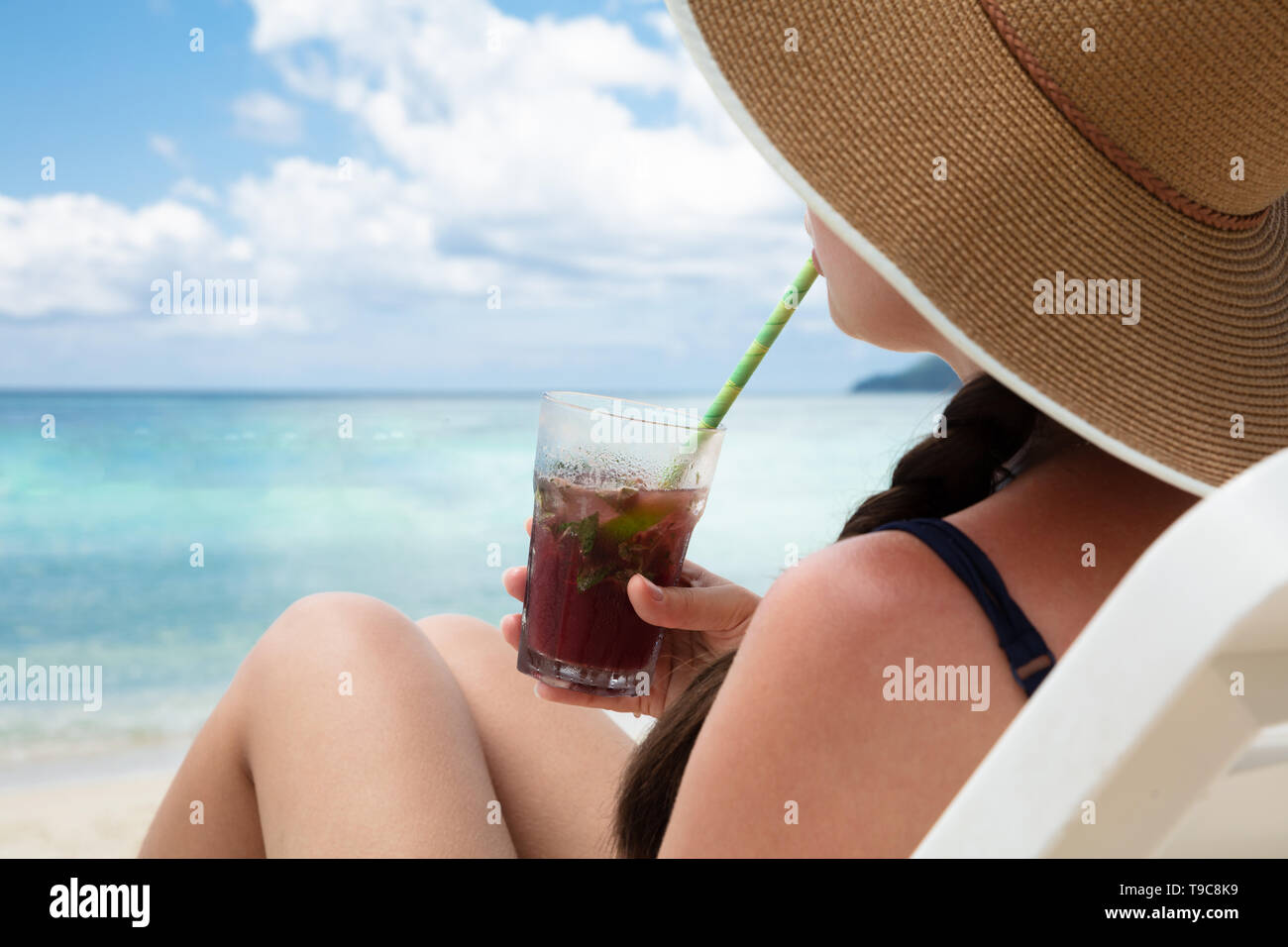Woman Sitting On Deck Chair Wearing Hat Drinking The Juice With Straw At The Beach Stock Photo