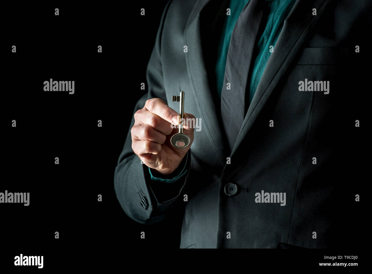 Close-up of incognito man in black suite, holding vintage key with two fingers, standing isolated on black background. Stock Photo
