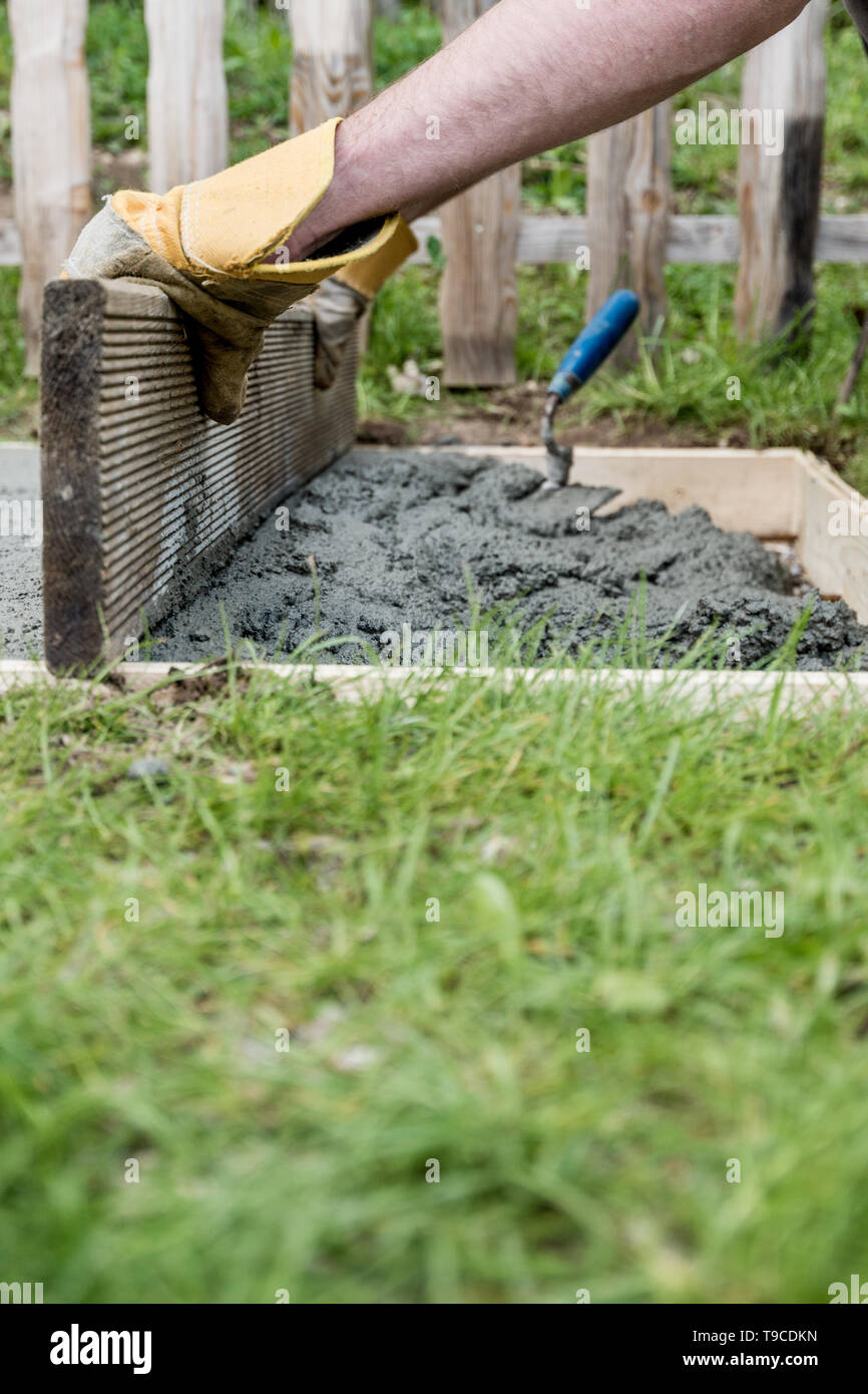 Low angle view of a man wearing protective work gloves leveling fresh cement as he makes a foundation in backyard. Stock Photo