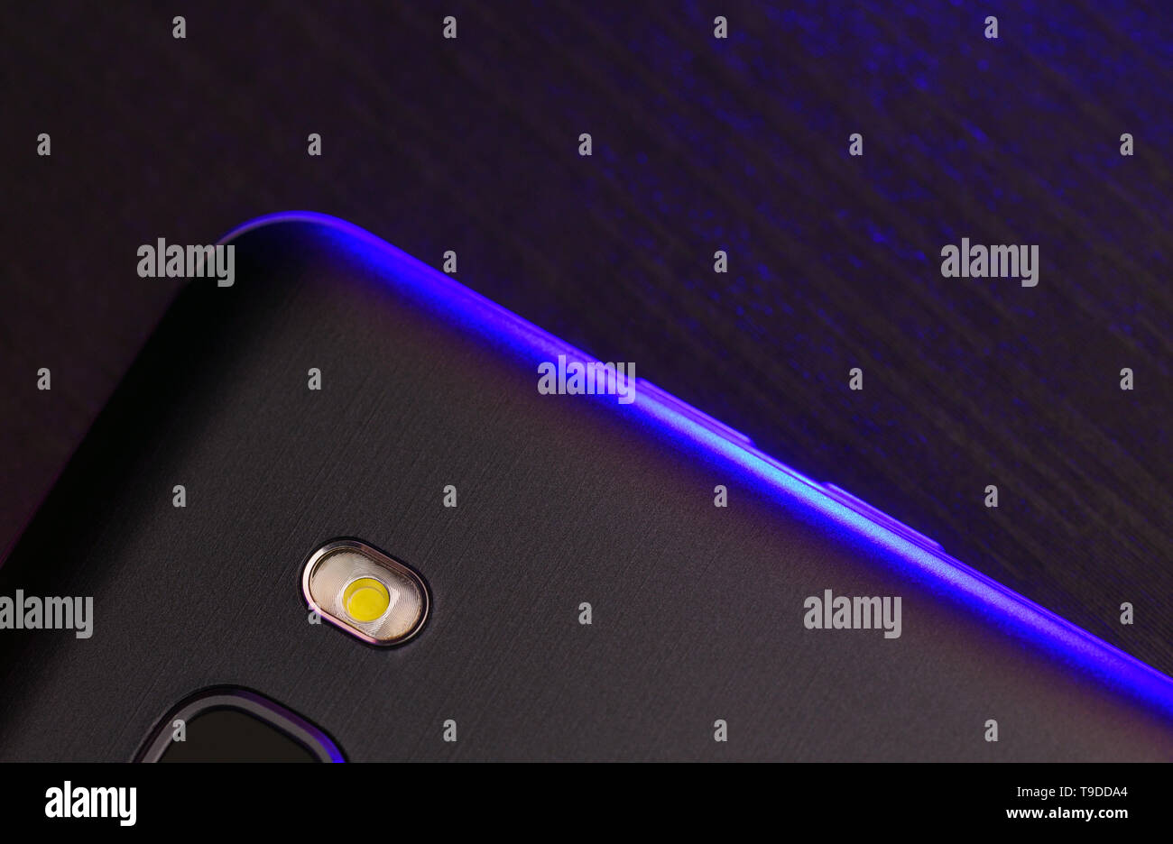 A fragment of the cell phone case in black with blue backlight. The phone lies on the black technological surface of the camera up with blue neon ligh Stock Photo