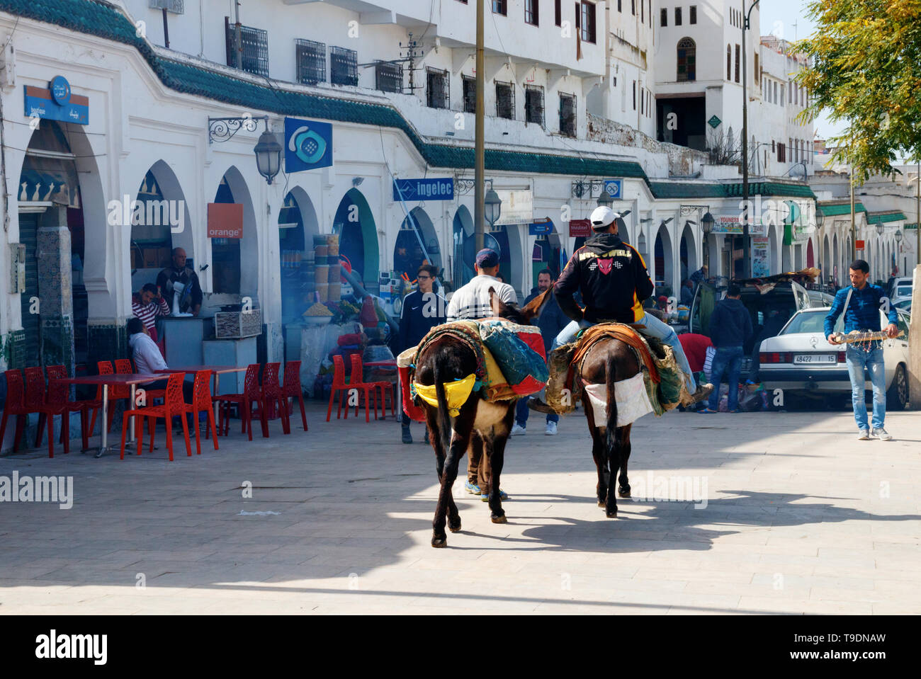 Main square of Moulay Idriss with shops, bars and two men with donkeys walking along the arcade on a sunny day. Moulay Idriss Zerhoun, Morocco Stock Photo