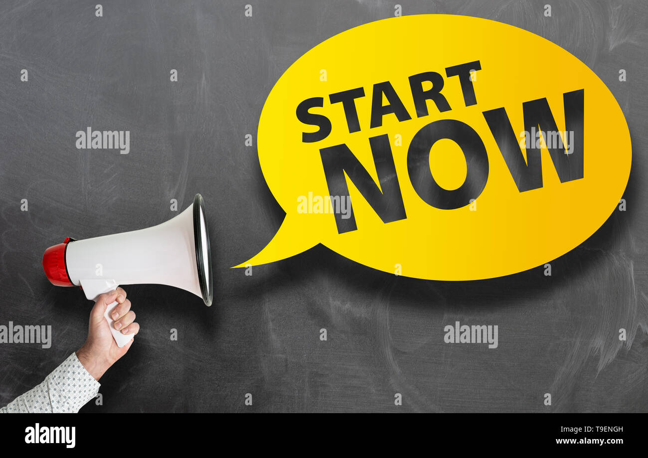 text START NOW in speech bubble and hand holding megaphone against blackboard Stock Photo