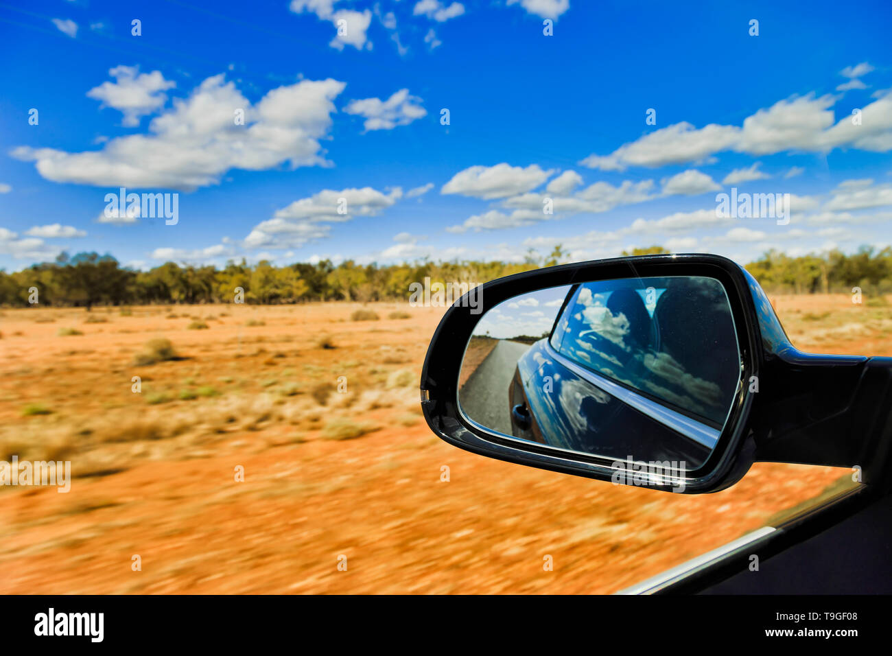 Australian outback red soil and scarse gum-trees under blue sky with rear view mirror through driving car blurring road side. Stock Photo