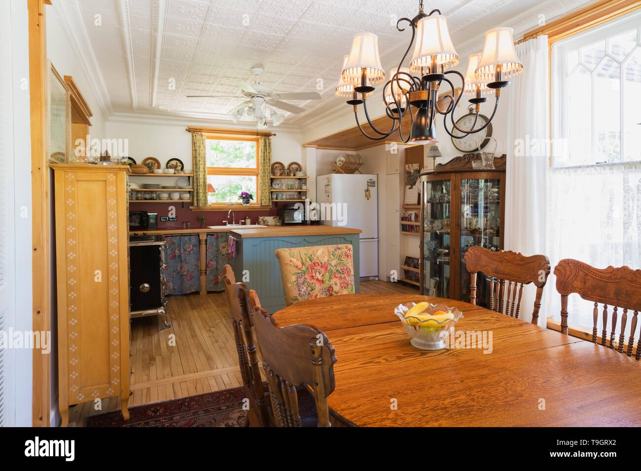 Oval oak wood dining table with press-back chairs, pinewood and glass  cabinet in dining room plus view of kitchen with turquoise painted pinewood  and maple wood top island in background inside an