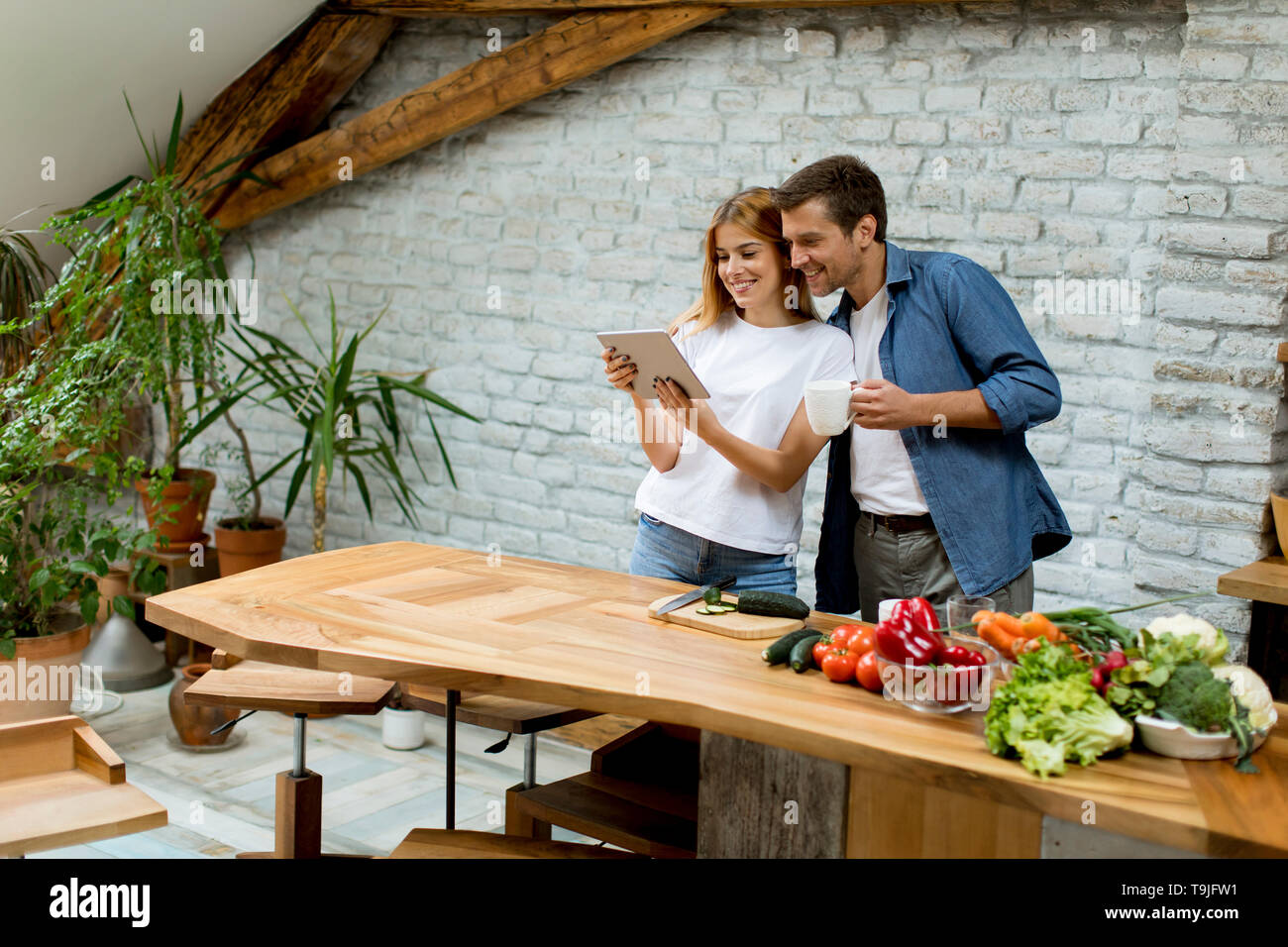 Lovely cheerful young couple cooking dinner together, looking recipe at digital tablet and having fun at rustic kitchen Stock Photo