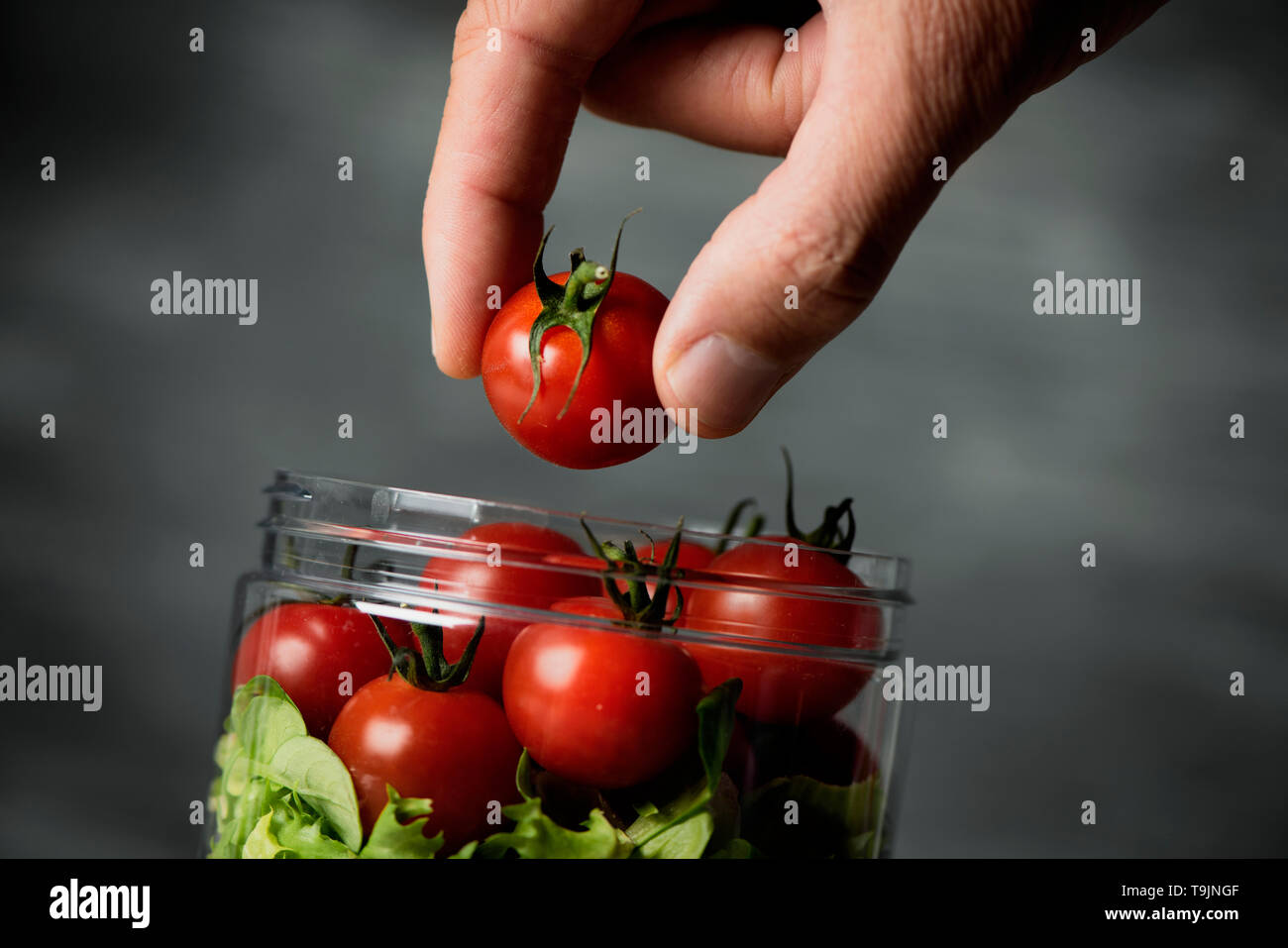closeup of a young caucasian man preparing or eating a jar salad, with cherry tomatoes and a mix of different leaf vegetables, against a gray backgrou Stock Photo
