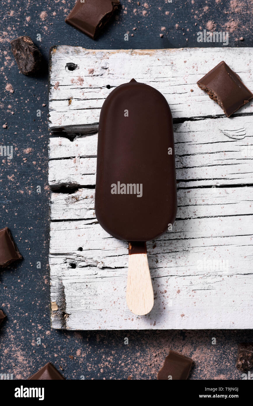 high angle view of a chocolate ice cream bar on a white rustic piece of wood, placed on a dark rustic wooden table Stock Photo