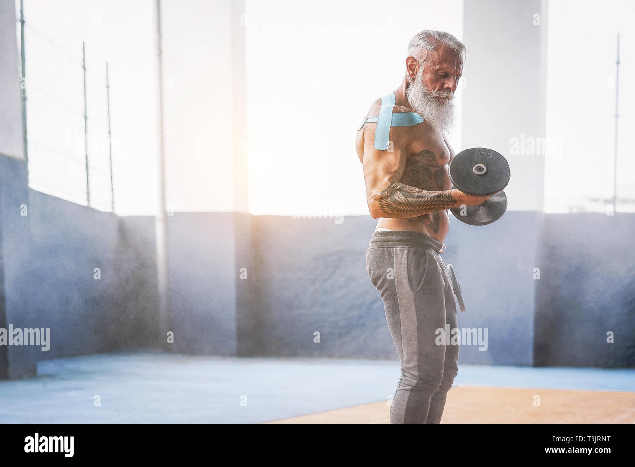 Fitness beard man doing biceps curl exercise inside a gym - Tattoo senior man training with dumbbells in wellness club center Stock Photo