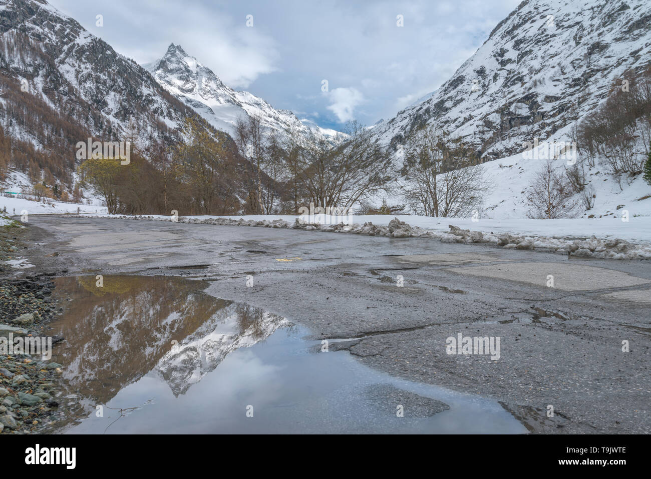 Prominent peak reflected on a still water puddle on the road. Sharp snowcapped peak reflections on a pool, perfect mirror image of a mountain. Stock Photo