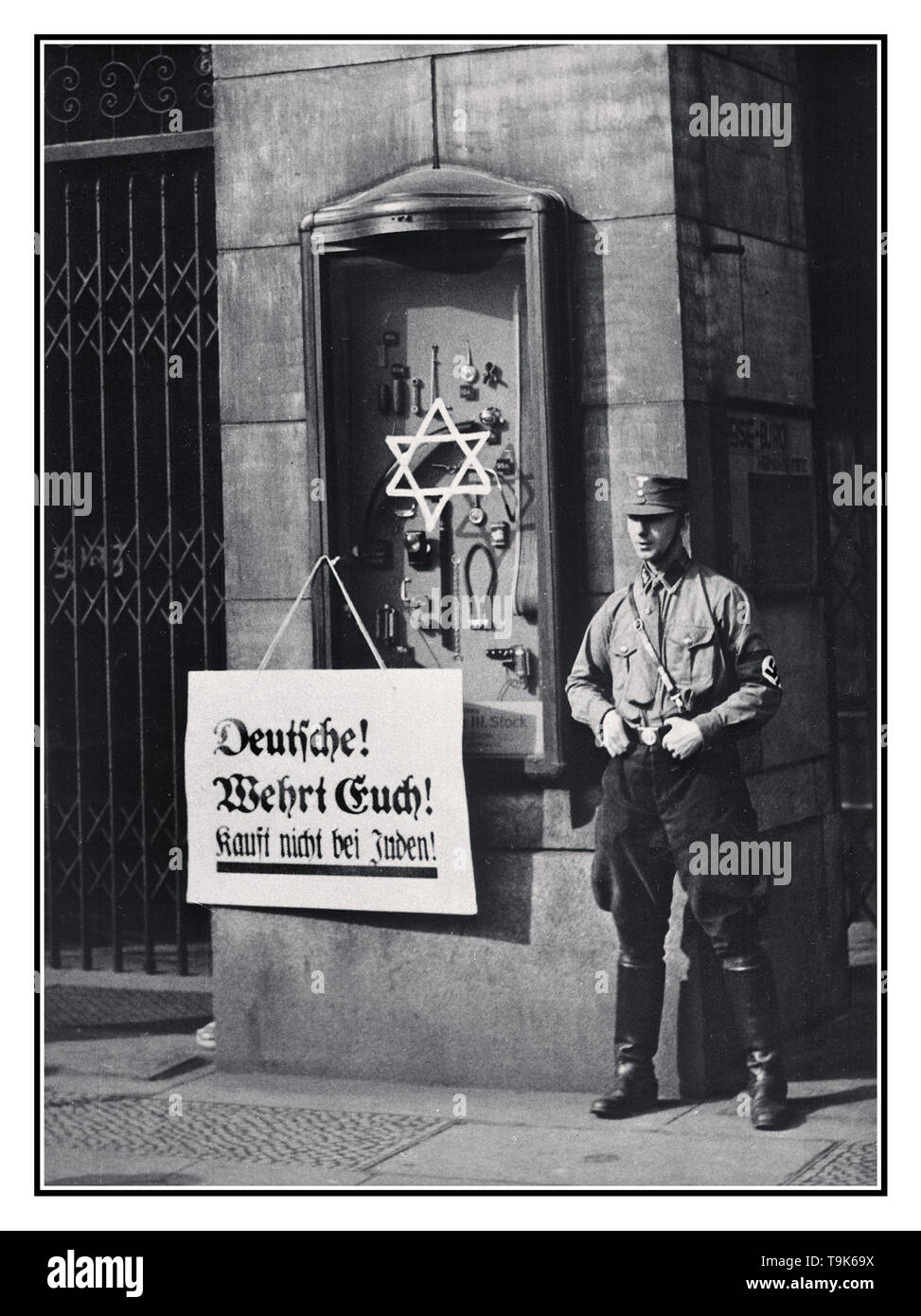 1930’s Germany Nazi boycott Jewish owned shops Vintage archive news propaganda anti-semitic image illustrating the April 1st,1933 Jewish shop store buying boycott which was announced by the Nazi NSDAP National Socialist Party. Placard reads, 'Germans, defend yourselves, do not buy from Jews', at the Jewish Tietz owned store, with Star of David stuck on a promotional cabinet. A Nazi NSDAP party member in uniform with swastika armband and wearing jackboots, stands outside to enforce the official state sponsored recommended boycott Stock Photo
