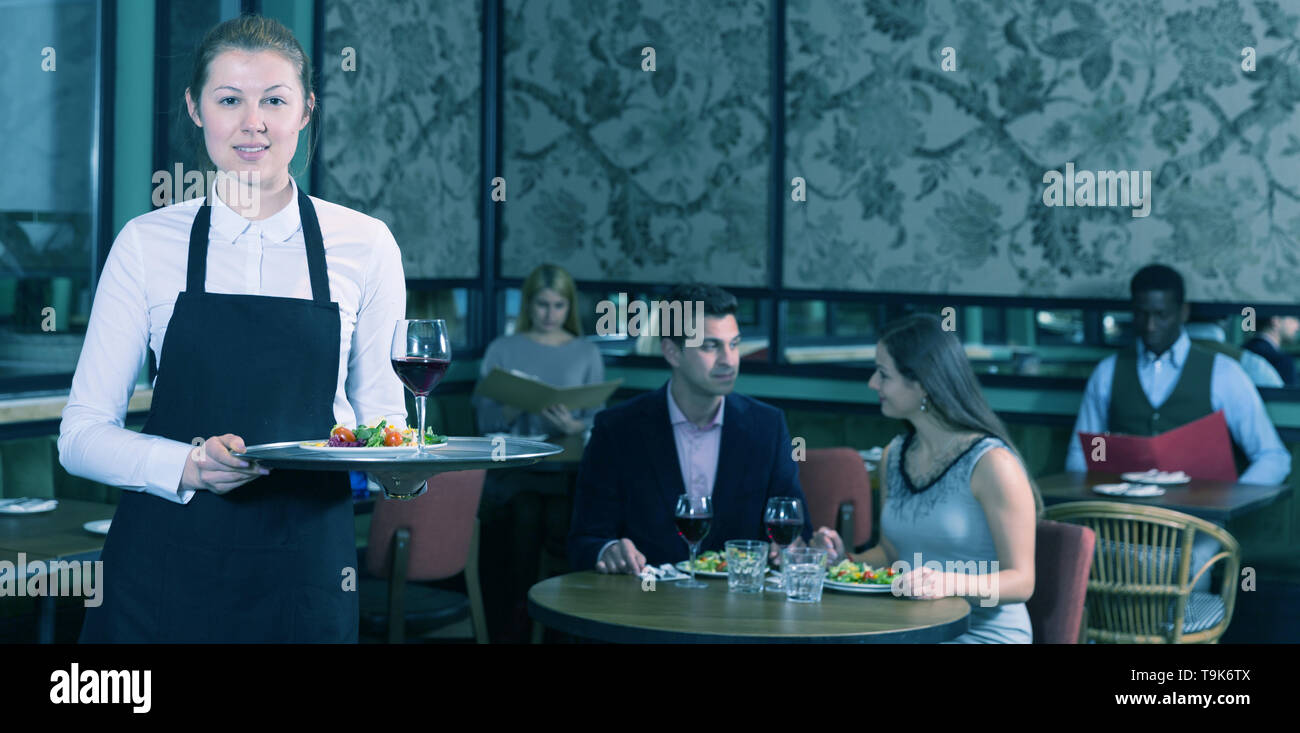 Polite waitress wearing apron holding tray with dishes in restaurant ...