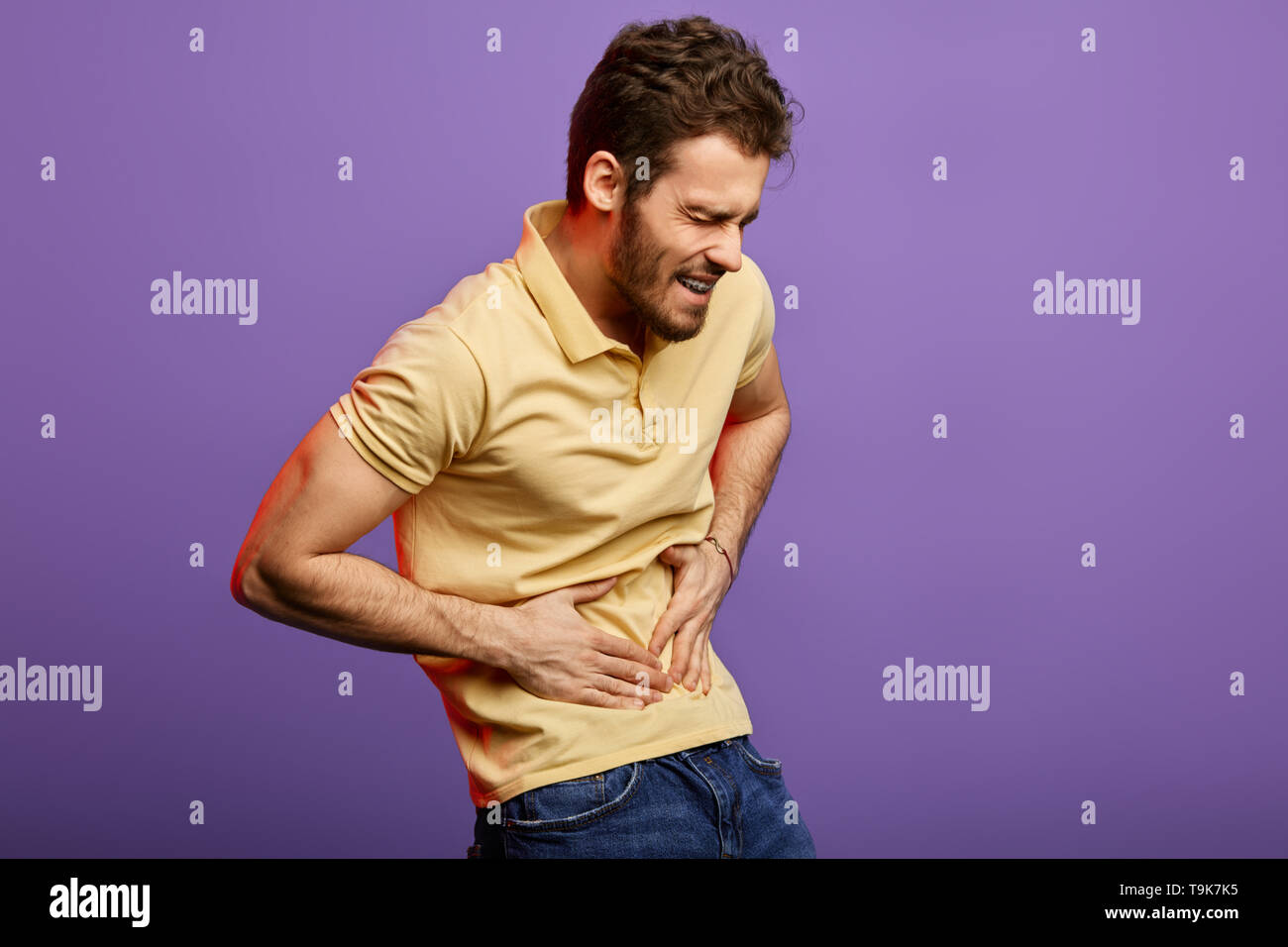 angry weak man having problems with stomach. student suffering from gastritis, gastroparesis, diarhea, Crohn's disease, cancer. isolated blue background. studio shot. man with awful pain in stomach Stock Photo