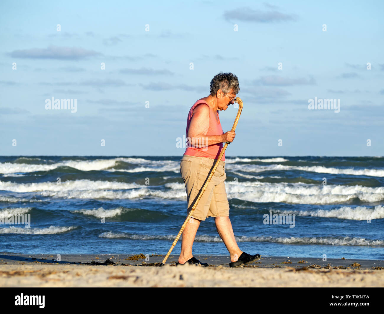 An elderly Caucasian woman with a walking stick strolls beside the Gulf Of Mexico on the beach during golden hour in Port Aransas, Texas USA. Stock Photo