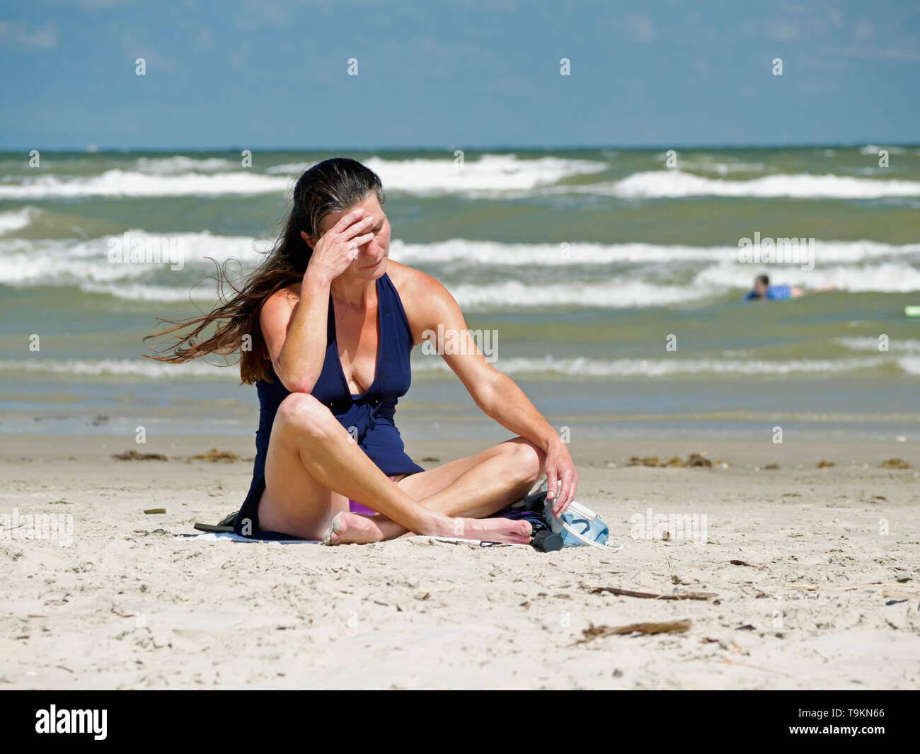 A brunette Caucasian woman sits cross legged on the beach with her hand to her forehead as if not feeling well, perhaps suffering from a headache. Stock Photo