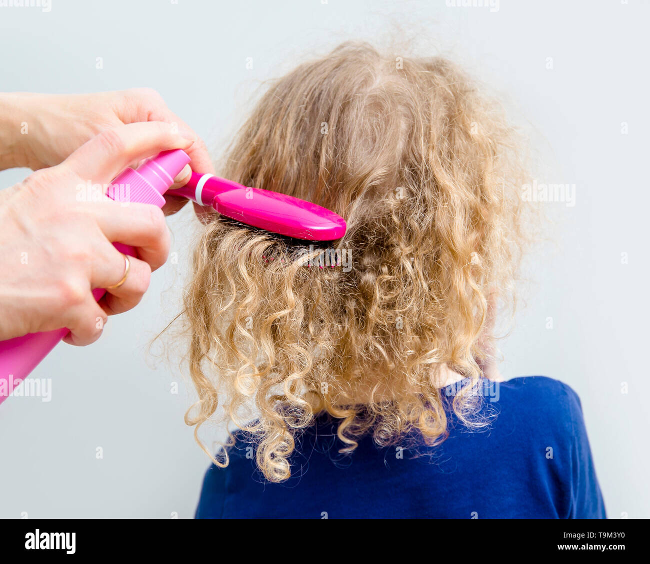 Close up view of mother hands spray curly hair balm on to girl child hair to help combing messy curly hair concept. Indoors minimal gray background. Stock Photo