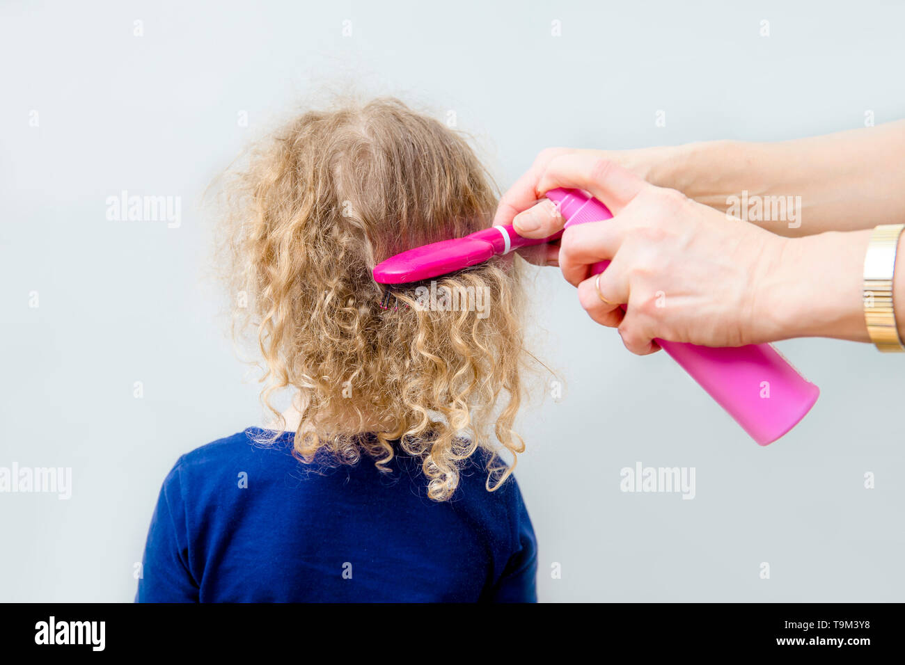 Close up view of mother hands spray curly hair balm on to girl child hair to help combing messy curly hair concept. Indoors minimal gray background. Stock Photo