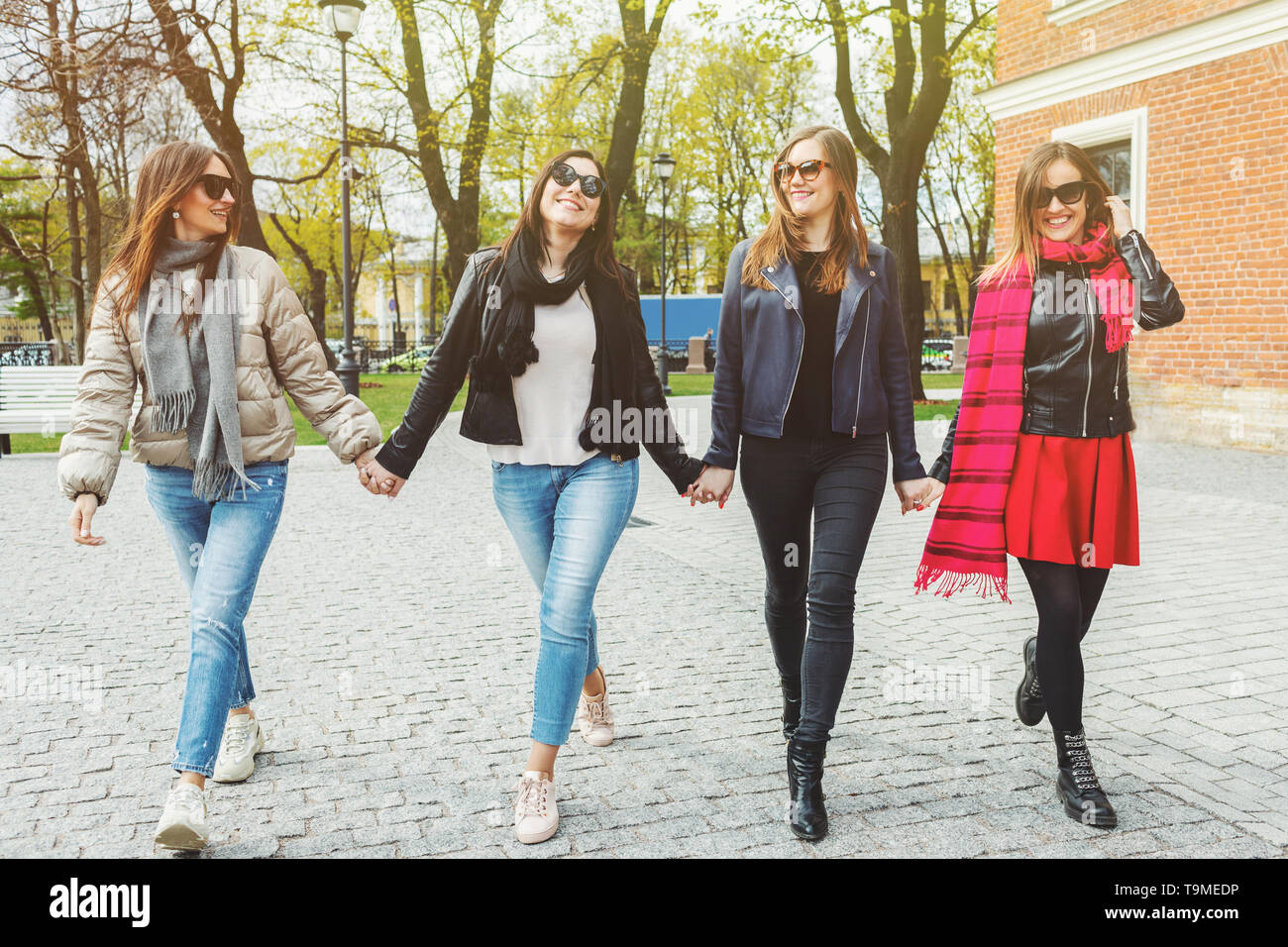 Four cheerful and beautiful women walk, holding hands, on the spring city in sunglasses. The concept of female friendship and power. Stock Photo