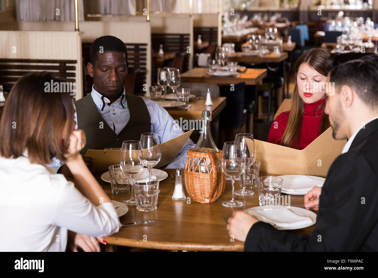 Group of young people sitting at table in cozy restaurant, choosing dishes from menu Stock Photo