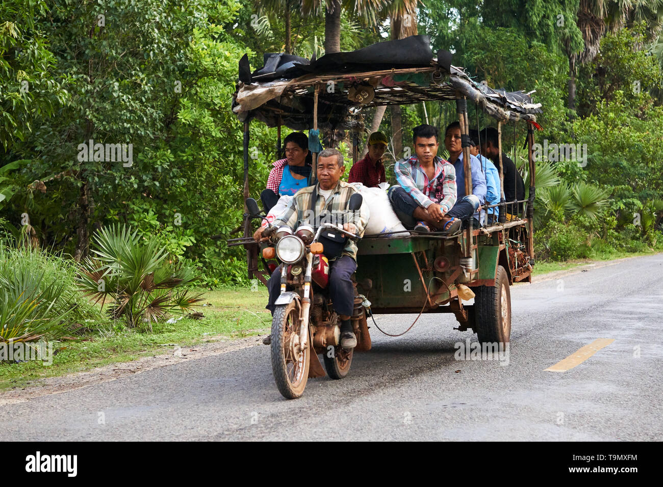 A mature Cambodian man drives a motorcycle while pulling a trailer (remorque) full of many adult passengers. Stock Photo