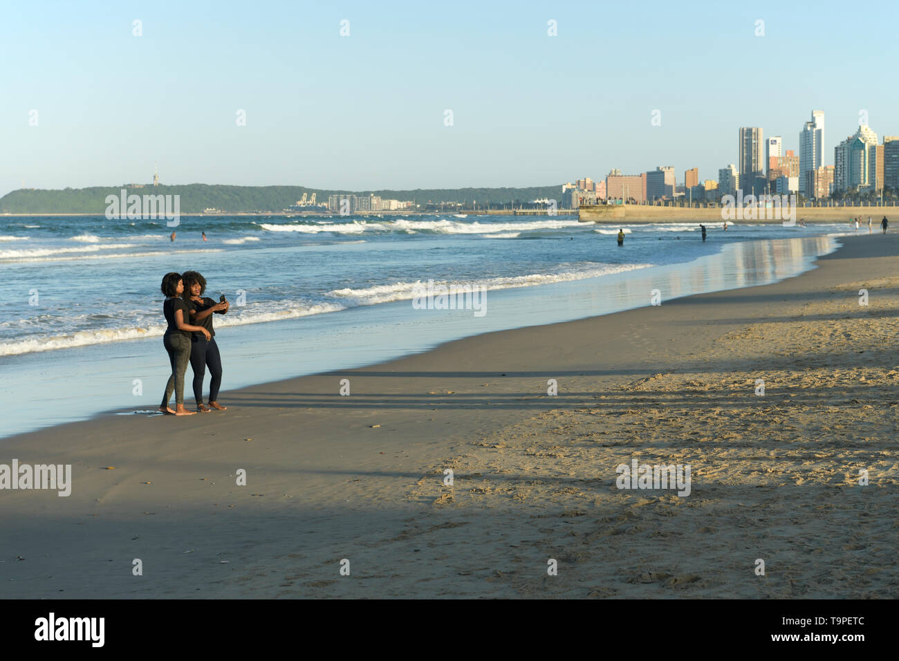 Two young adult women friends taking selfie with mobile phone, standing on beach, Durban, KwaZulu-Natal, South Africa, people, ethnic, landscape, city Stock Photo