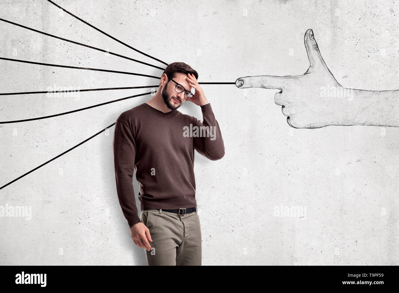 Handsome man in casual clothes standing in half-turn, with hand on forehead, near wall with drawing of finger gun shooting at him. Stock Photo