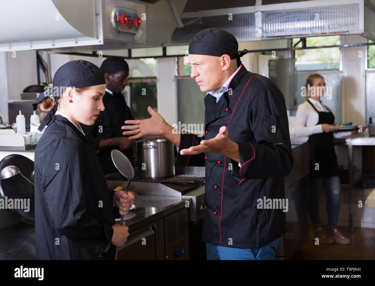 Enraged chef expressing dissatisfaction with work of frustrated girl in restaurant kitchen Stock Photo