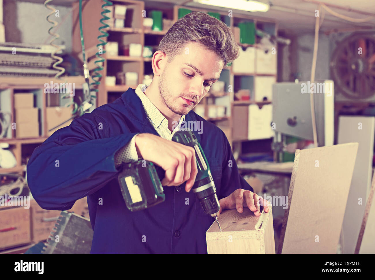 Young smiling joiner using drilling machine to make drawer at workshop Stock Photo