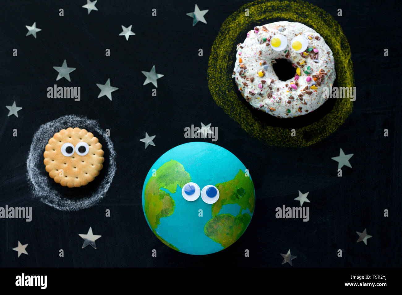cute model of the Earth, Sun and Moon with googly eyes on the chalkboard, a donut, a globe and a cookie on the chalkboard Stock Photo
