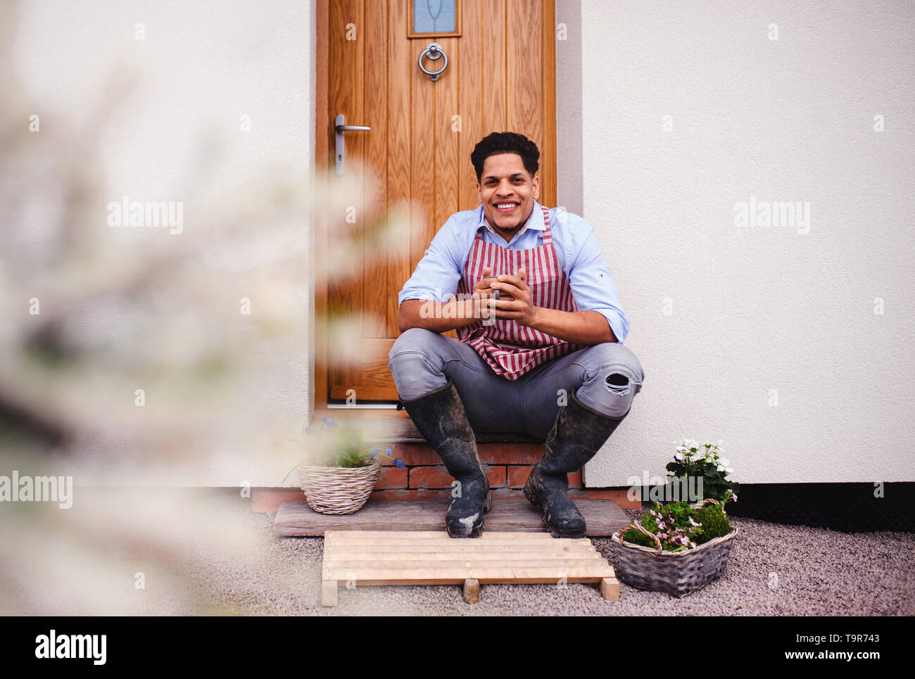 A portrait of young man gardener sitting in front of door at home. Stock Photo